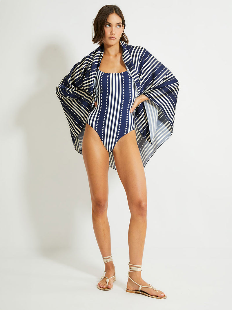 woman standing wearing lemlem ADIA Sarong featuring a classic navy background with ivory stripes and dots on a silky fabric and a matching one piece swimsuit