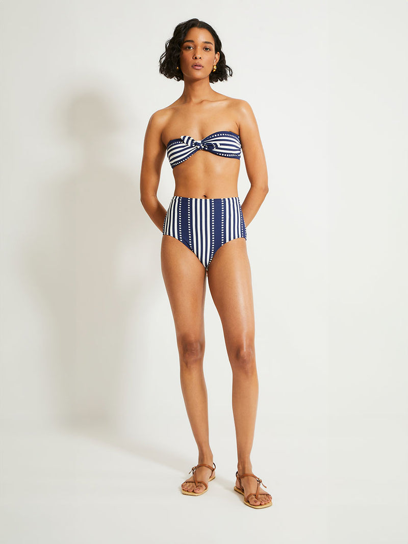 Woman Standing Wearing lemlem ELSI High Waist Bottom featuring a classic navy background with ivory stripes and dots and matching bandeau top