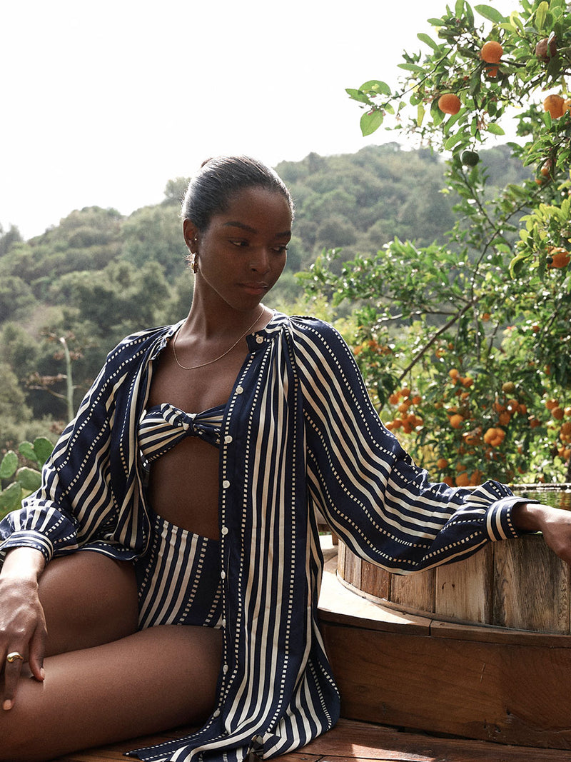 Woman Sitting by the pool in a tree house wearing lemlem bandeau bikini top and high waist bottom featuring stripe and dots pattern in white color on navy background and matching meaza button up dress