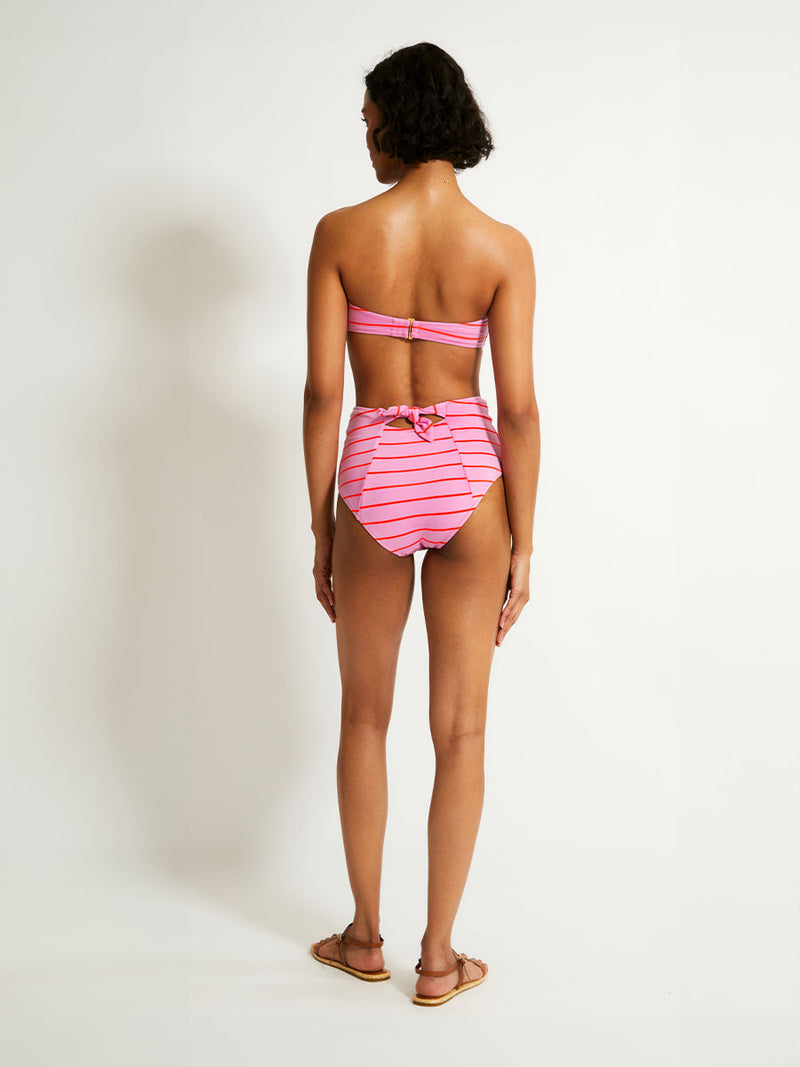 Back of a Woman Standing Wearing lemlem AVA Bandeau Top featuring soft pink color accented by neon orange red stripes and matching high waist bottom