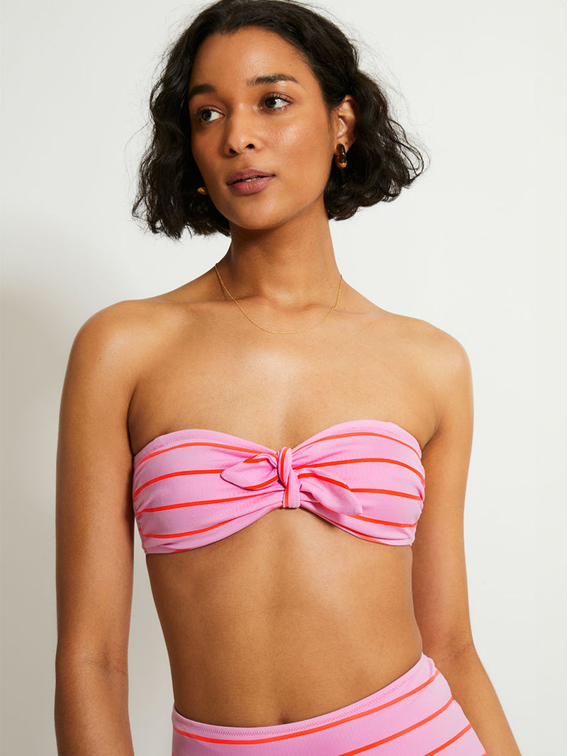 Woman Standing Wearing lemlem AVA Bandeau Top featuring soft pink color accented by neon orange red stripes and matching high waist bottom