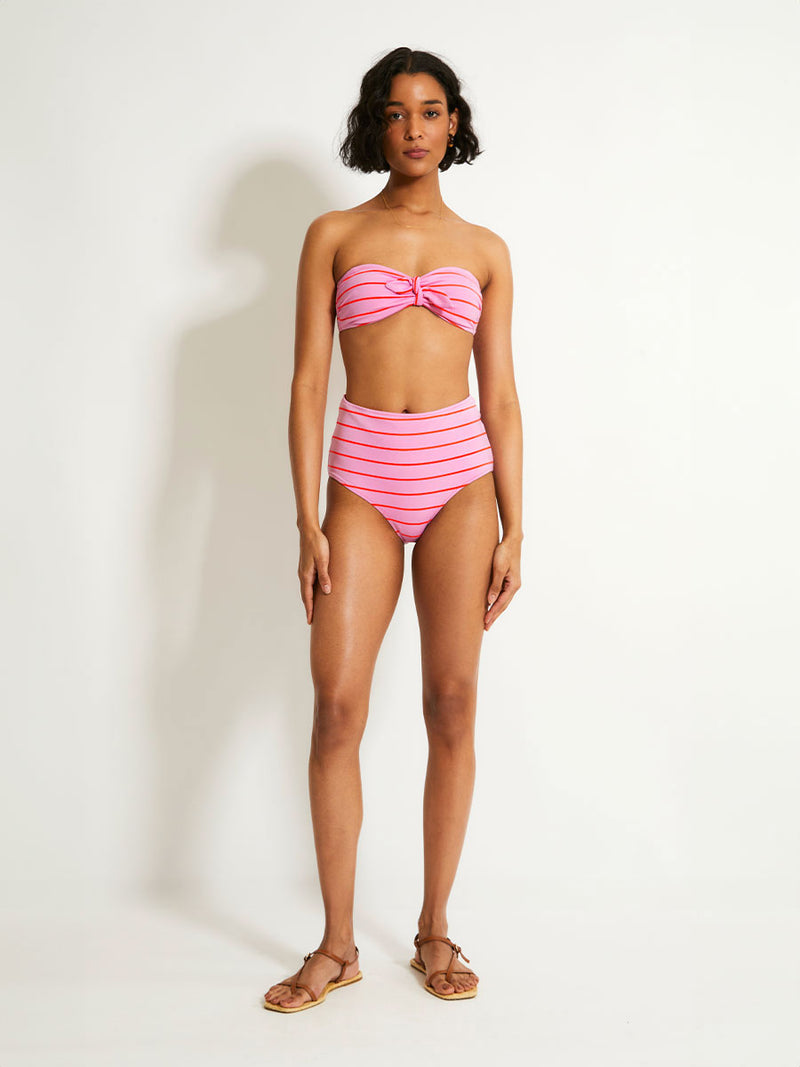 Woman Standing Wearing lemlem AVA Bandeau Top featuring soft pink color accented by neon orange red stripes and matching high waist bottom