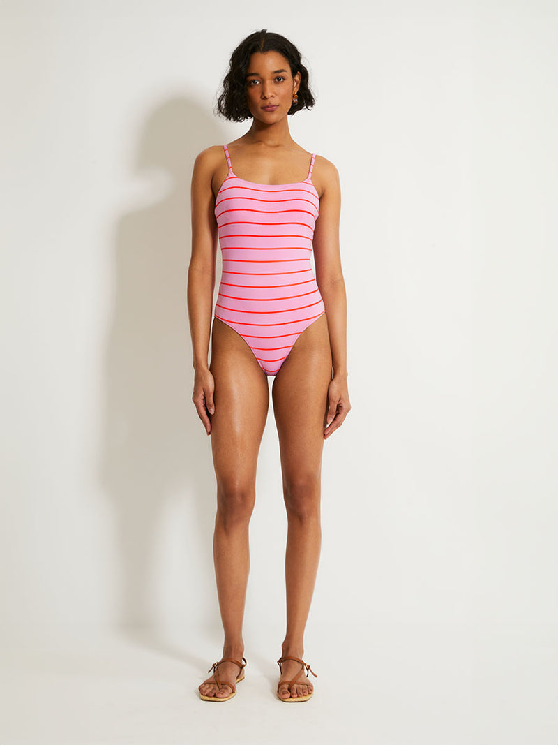 Woman Standing Wearing lemlem ELENE One Piece featuring  soft pink color accented by neon orange red stripes 