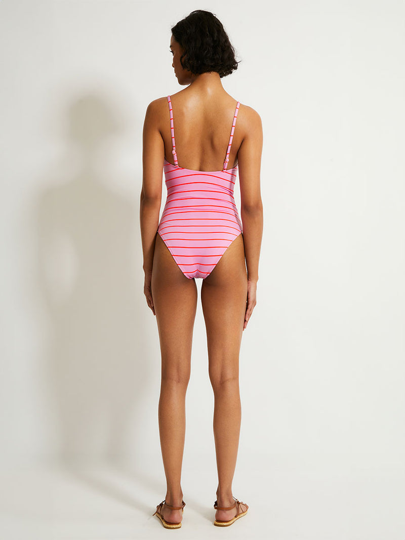 Back of a Woman Standing Wearing lemlem ELENE One Piece featuring  soft pink color accented by neon orange red stripes 