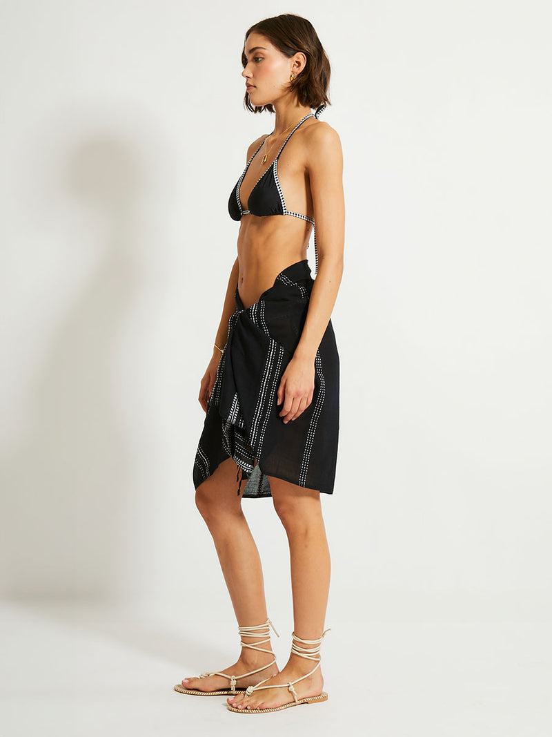 Side shot of a Woman Standing Wearing lemlem LEMA Sarong featuring classic back color with white dotted lines and black triangle top