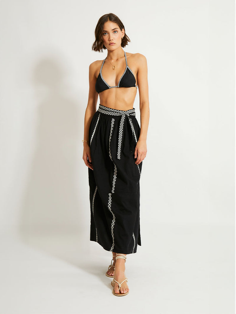 Woman Standing Wearing lemlem TOLA Skirt Featuring rich black background with intricate cream Tibeb border pattern and black triangle bikini top