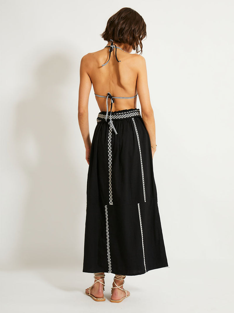 Back of a Woman Standing Wearing lemlem TOLA Skirt Featuring rich black background with intricate cream Tibeb border pattern and black triangle bikini top