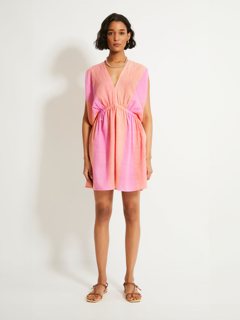 Woman Standing Wearing lemlem ALEM Plunge Dress featuring light, gauzy fabric with a beautiful ombre color block of pink, blush, and peach colors.