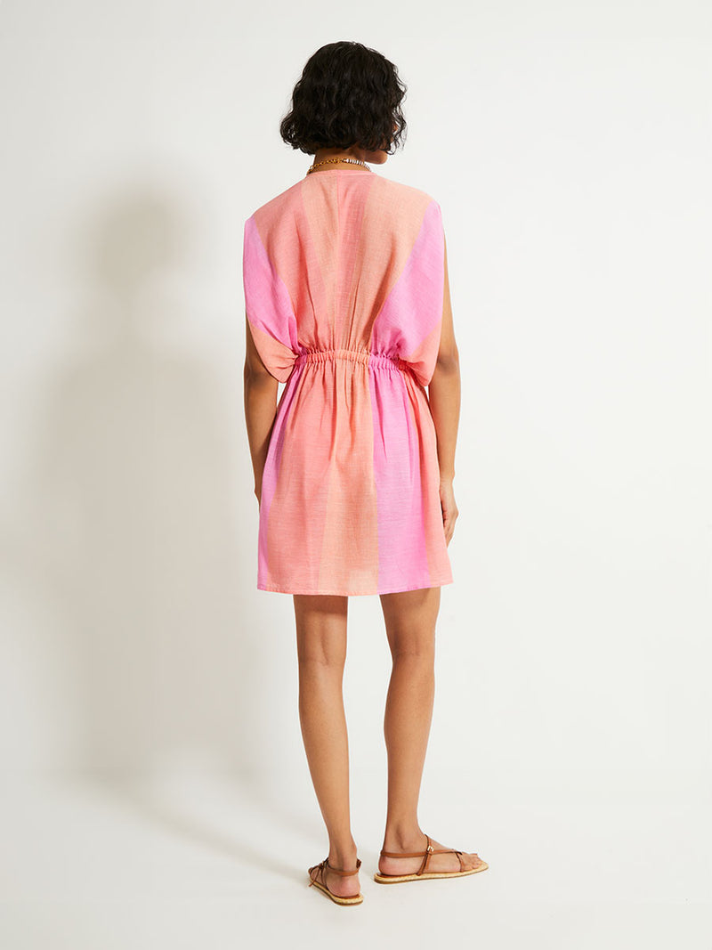 Back of a Woman Standing Wearing lemlem ALEM Plunge Dress featuring light, gauzy fabric with a beautiful ombre color block of pink, blush, and peach colors.