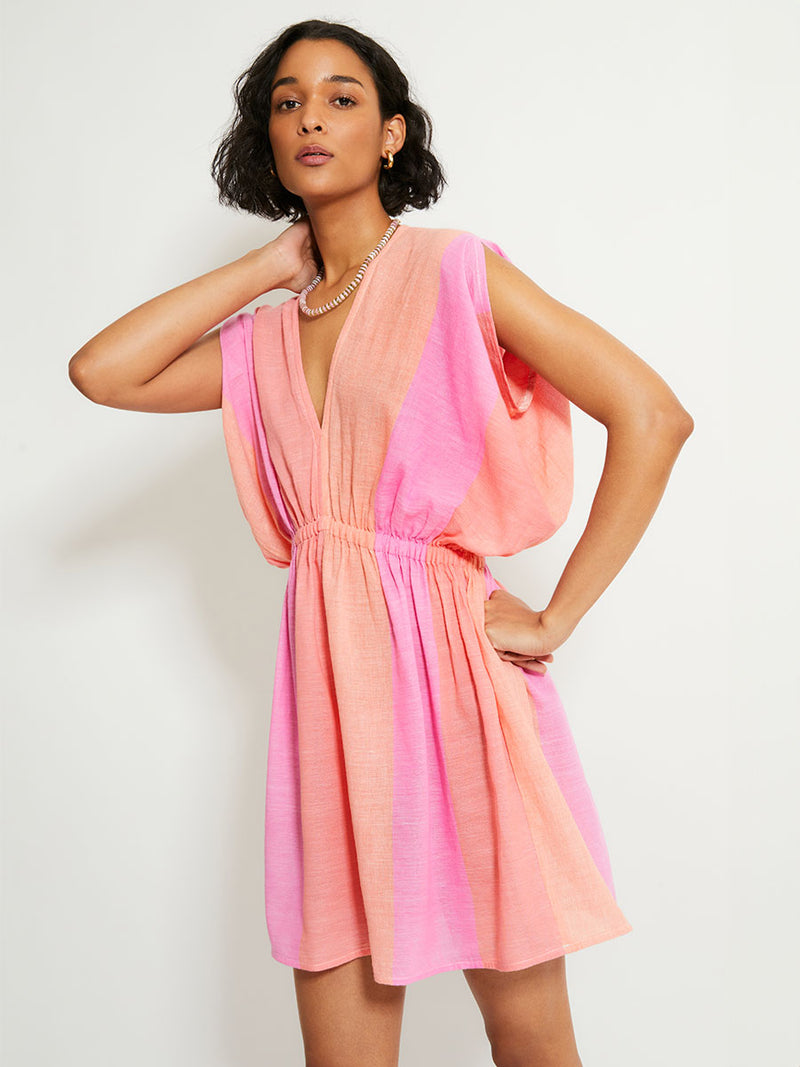 Woman Standing Wearing lemlem ALEM Plunge Dress featuring light, gauzy fabric with a beautiful ombre color block of pink, blush, and peach colors.