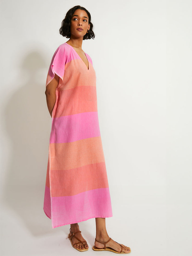 Side of a Woman Standing Wearing lemlem DALILA Caftan featuring gauzy fabric with a beautiful ombre color block of pink, blush, and peach colors.