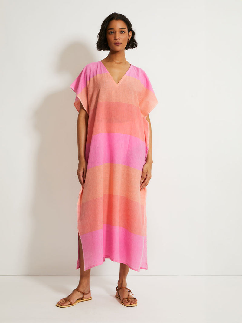 Woman Standing Wearing lemlem DALILA Caftan featuring gauzy fabric with a beautiful ombre color block of pink, blush, and peach colors.