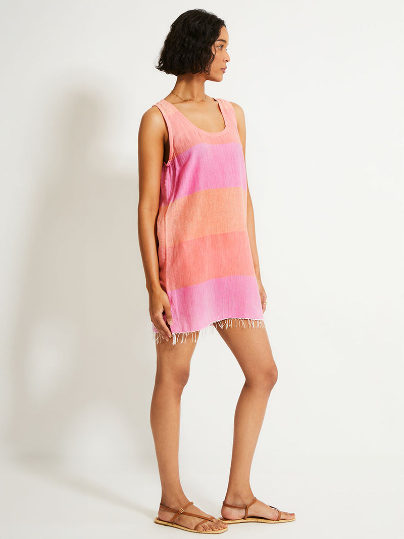 Side shot of a Woman Standing Wearing lemlem MAHLET Dress featuring gauzy fabric with a beautiful ombre color block of pink, blush, and peach colors.