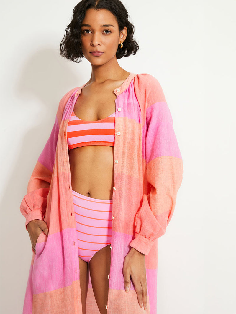 Woman Standing Wearing lemlem Makeda Dress featuring gauzy fabric with a beautiful ombre color block of pink, blush, and peach colors.