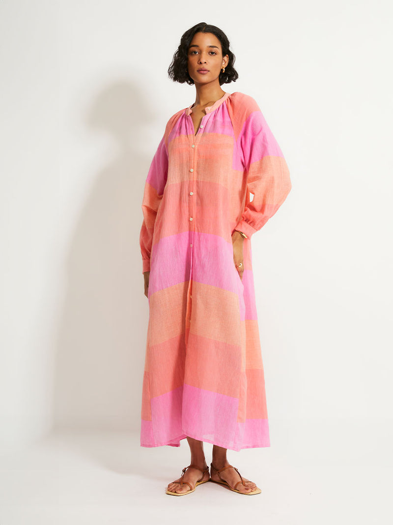 Woman Standing Wearing lemlem Makeda Dress featuring gauzy fabric with a beautiful ombre color block of pink, blush, and peach colors.