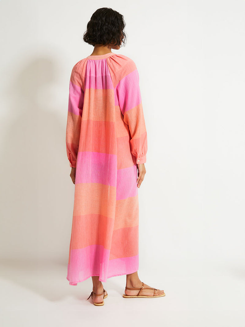 Back of a Woman Standing Wearing lemlem Makeda Dress featuring gauzy fabric with a beautiful ombre color block of pink, blush, and peach colors.