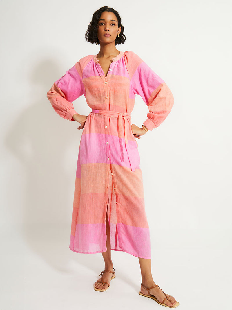 Woman Standing Wearing lemlem Makeda Dress featuring gauzy fabric with a beautiful ombre color block of pink, blush, and peach colors.