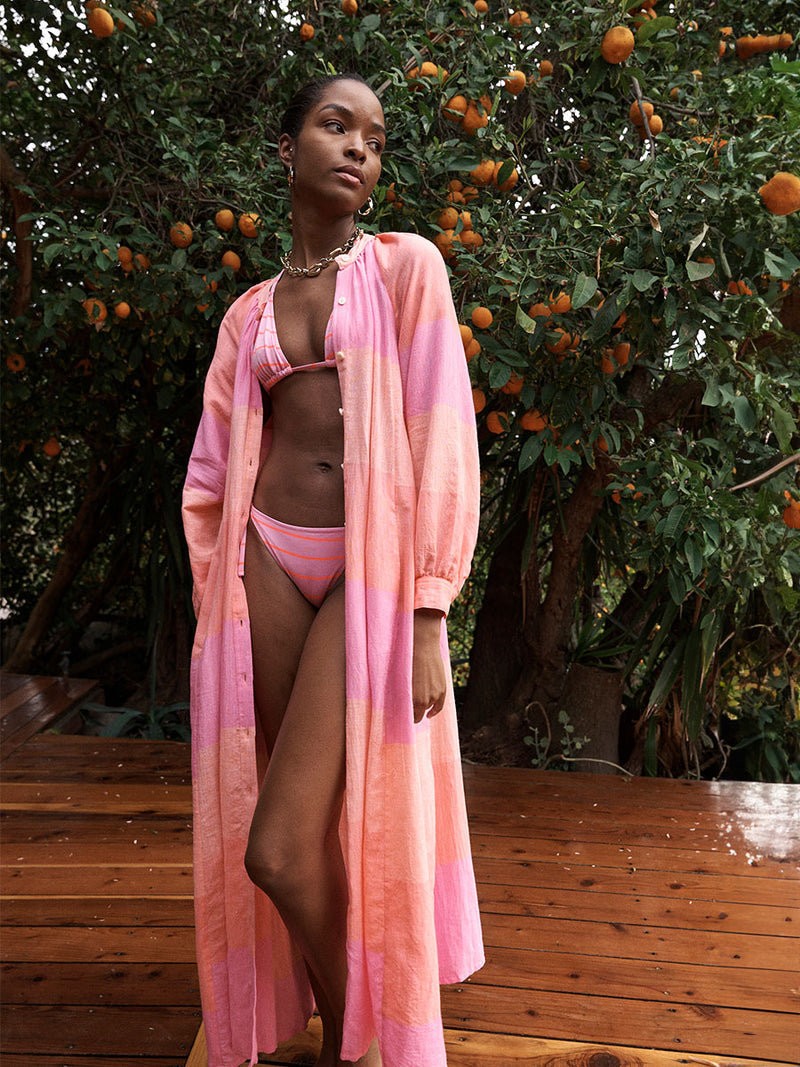 Woman Standing Wearing lemlem Makeda Dress featuring gauzy fabric with a beautiful ombre color block of pink, blush, and peach colors and bikini swimsuit featuring thin stripe pattern