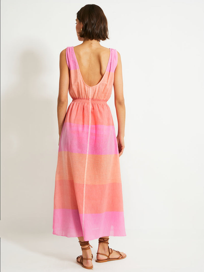 Back of a Woman Standing Wearing lemlem Melat Scoop Dress featuring gauzy fabric with a beautiful ombre color block of pink, blush, and peach colors.