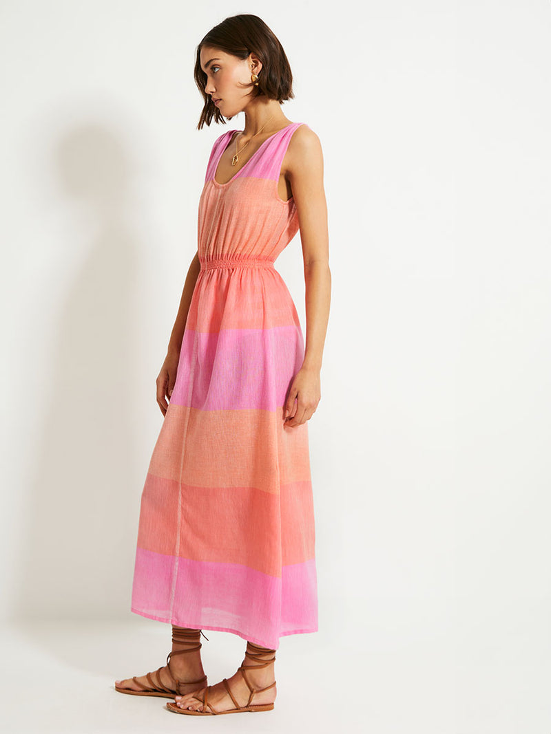 Side View of a Woman Standing Wearing lemlem Melat Scoop Dress featuring gauzy fabric with a beautiful ombre color block of pink, blush, and peach colors.