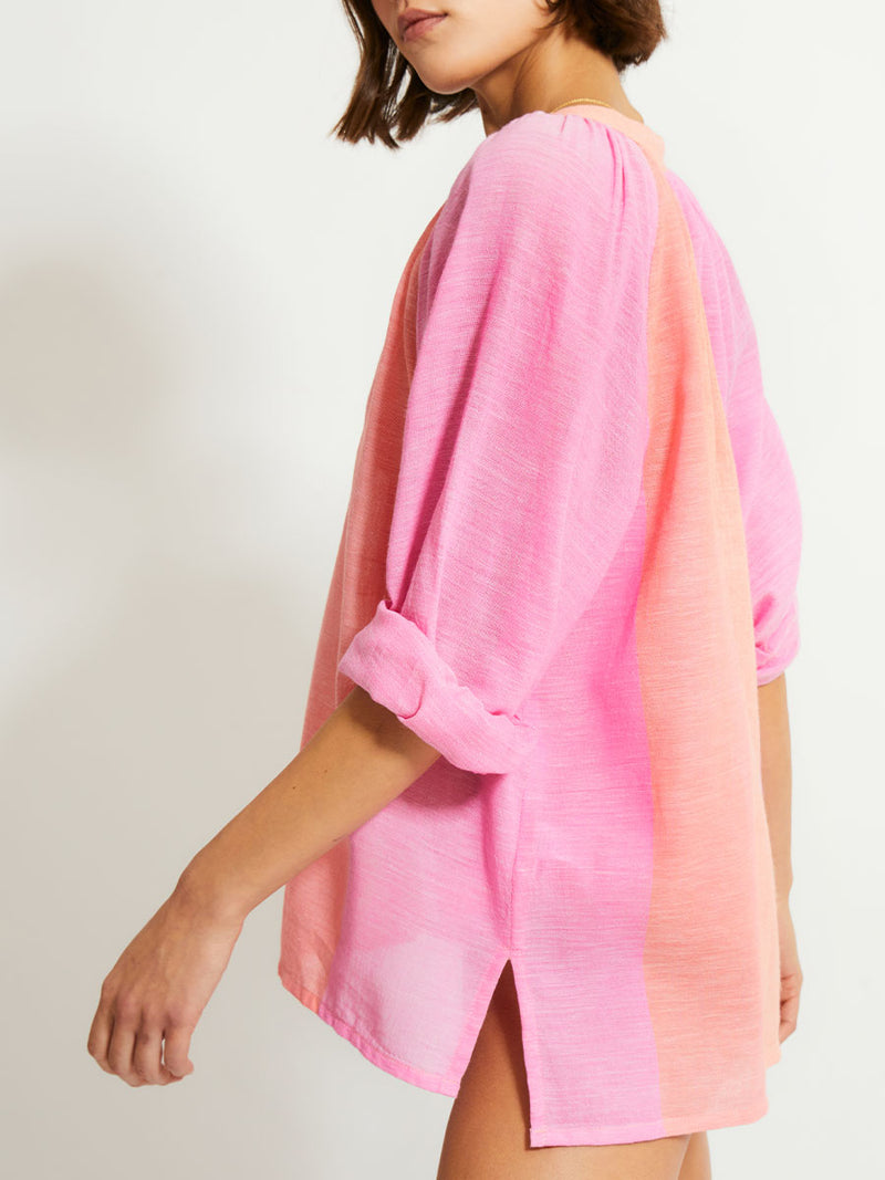 Side Shot of a Woman Standing Wearing lemlem MITA Blouse featuring gauzy fabric with a beautiful ombre color block of pink, blush, and peach colors and brief bikini bottom