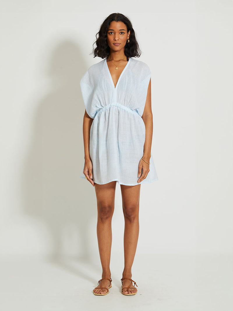 Woman Standing Wearing lemlem Alem Plunge Dress featuring airy gauze fabric in a delicate pale blue color.