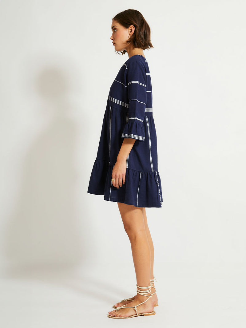 Side of a Woman Standing Wearing lemlem HANNA Flutter Dress in Nunu Navy featuring delicate white Tibeb bands on a rich navy background