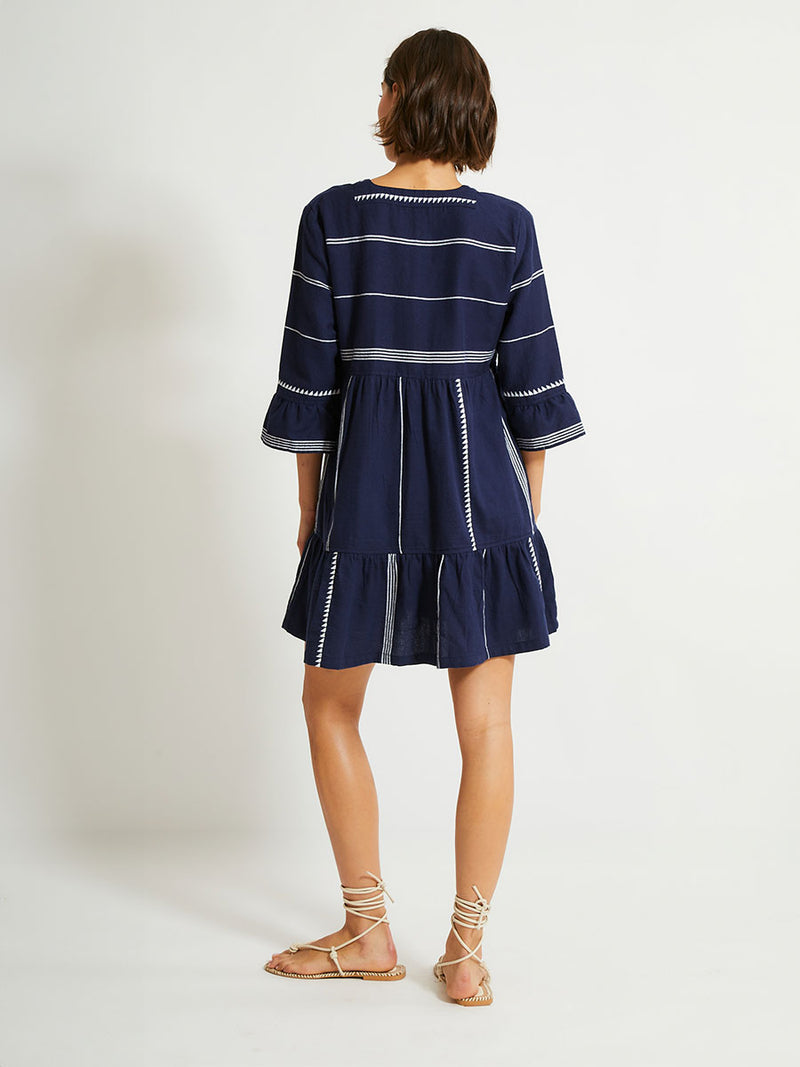 Back of a Woman Standing Wearing lemlem HANNA Flutter Dress in Nunu Navy featuring delicate white Tibeb bands on a rich navy background