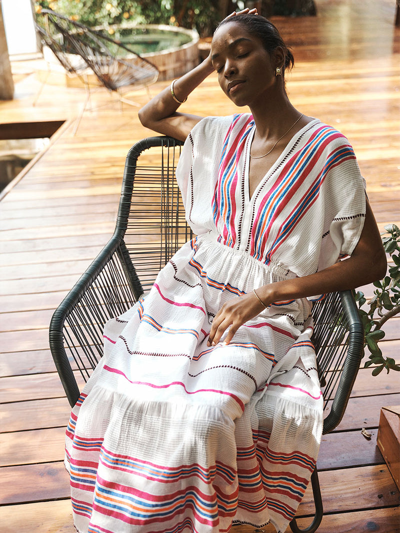 Woman Sitting on a straw chair  Wearing lemlem Leila Plunge Dress Featuring stripe pattern of bright magenta, blue, and orange on s crispy white background