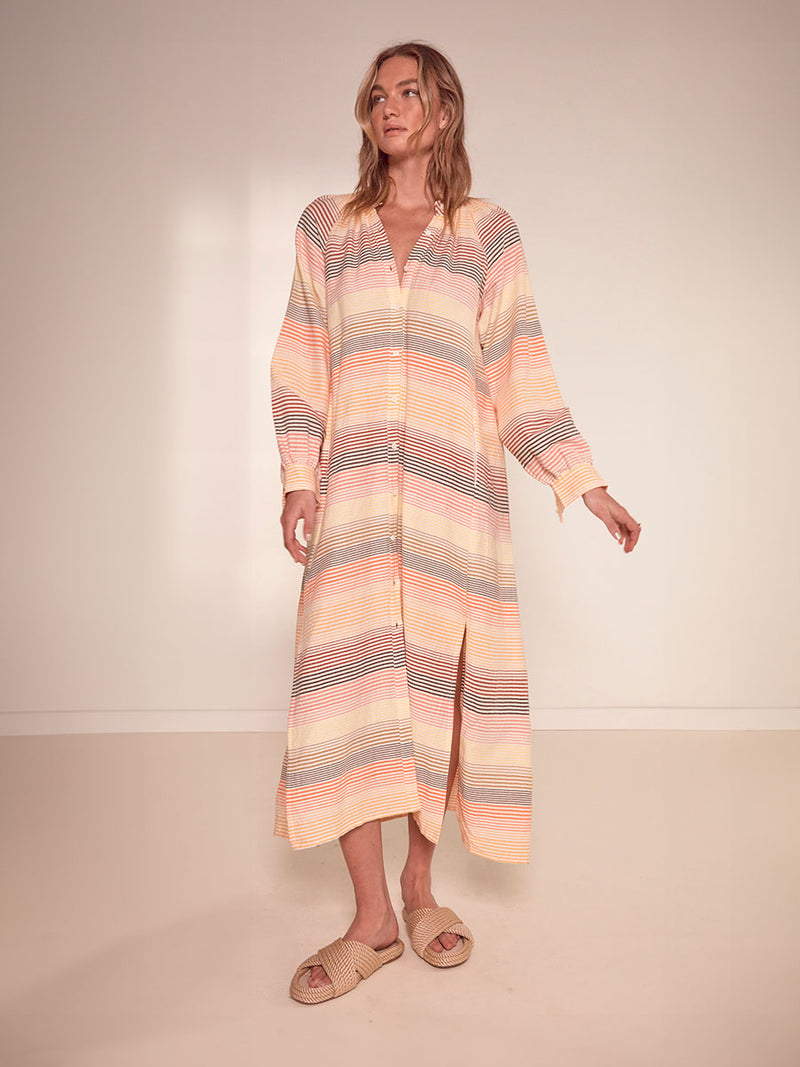 Woman Standing Wearing lemlem Makeda Button Up Dress Featuring continuous stripe pattern in warm yellow, orange and peach tones