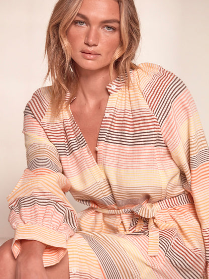 Close up on a Woman Sitting Wearing lemlem Makeda Button Up Dress Featuring continuous stripe pattern in warm yellow, orange and peach tones