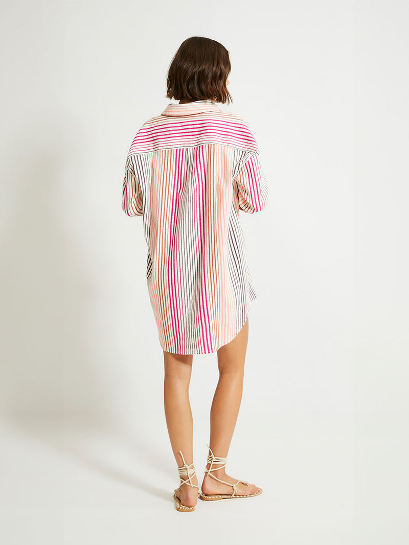 Back of a Woman Standing Wearing lemlem Mariam Shirt featuring ombre stripe pattern, where deep plum seamlessly transitions into peach, chestnut, and bright pink.