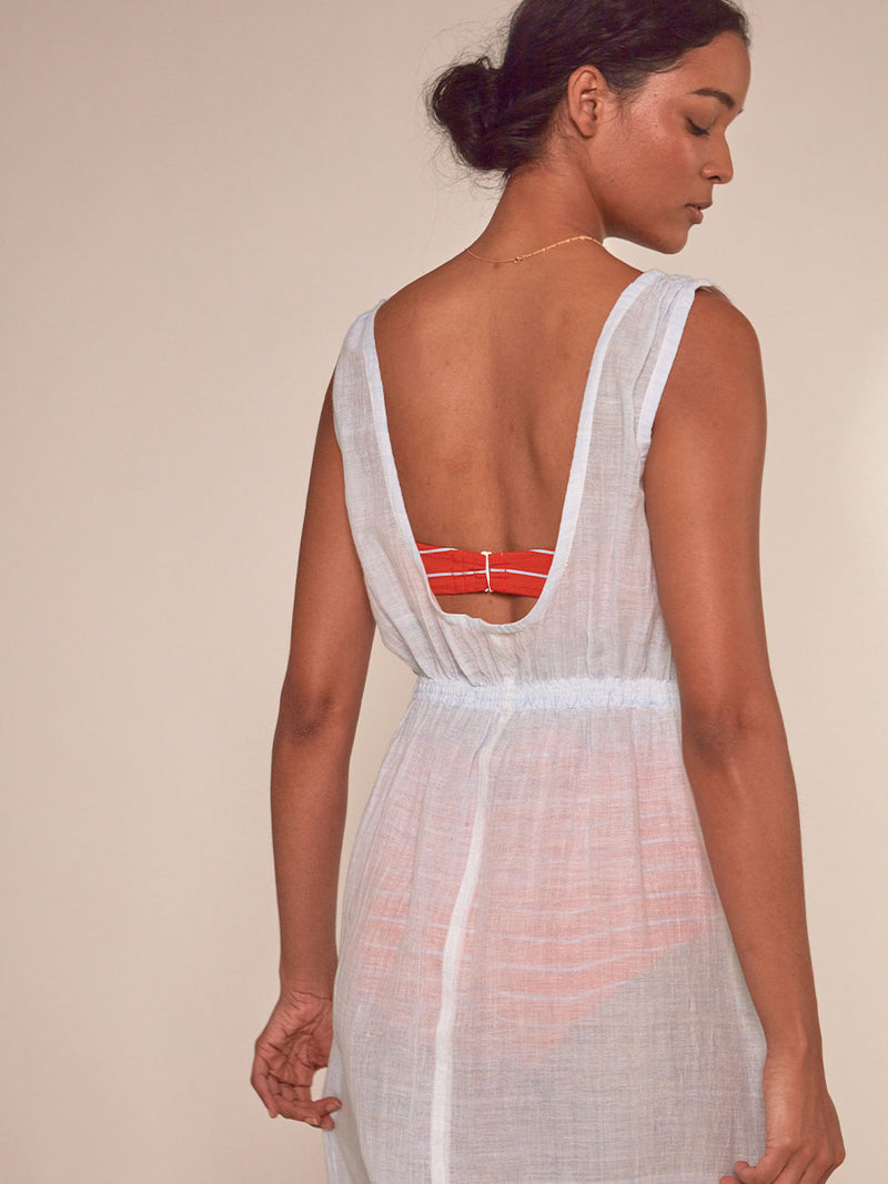 Back View of a Woman Standing Wearing lemlem Melat Scoop Neck Dress featuring airy gauze fabric in a delicate pale blue color, Jeliba Tangerine Bandeau  top and High Waist Bottom