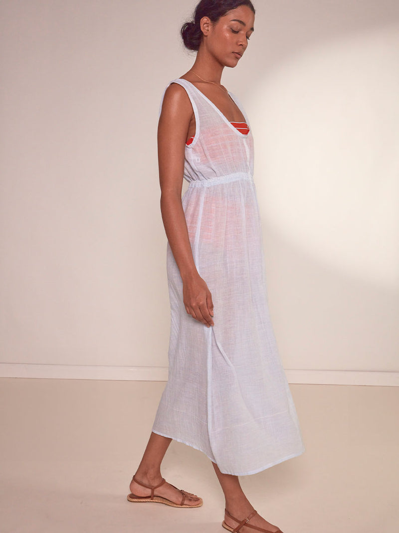Side View of a Woman Standing Wearing lemlem Melat Scoop Neck Dress featuring airy gauze fabric in a delicate pale blue color, Jeliba Tangerine Bandeau top and High Waist Bottom