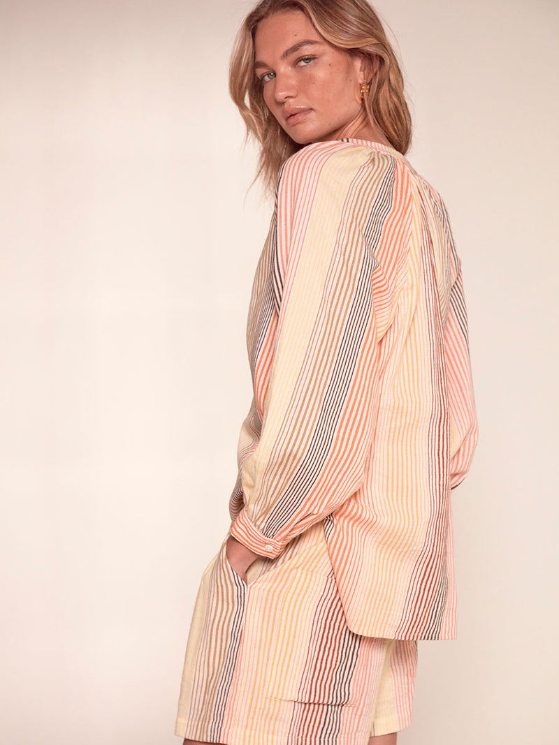 Side View of a Woman Standing Wearing lemlem Mita Button Up Blouse featuring continuous stripe pattern in warm yellow, orange and peach tones and matching Safia Shorts