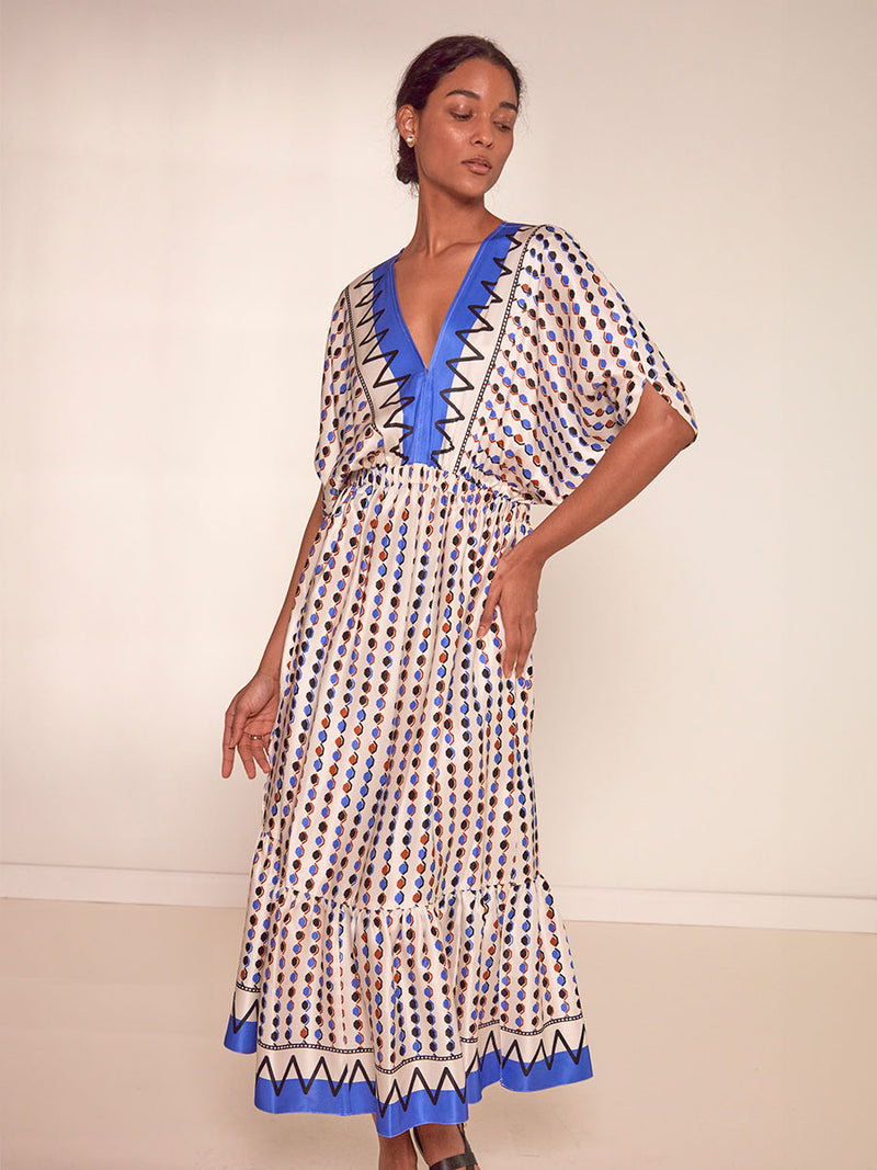 Woman standing wearing  lemlem Leila Plunge Dress featuring diamond pattern in natural terracotta and rich blue hues against a cream background