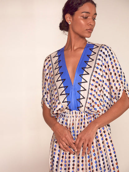 Close up on a Woman standing wearing lemlem Leila Plunge Dress featuring diamond pattern in natural terracotta and rich blue hues against a cream background
