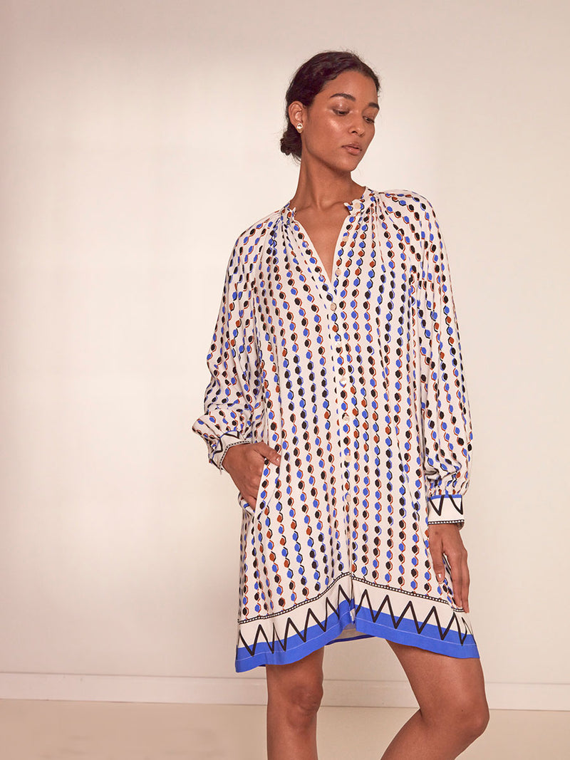  Woman Standing Wearing lemlem Meaza Button Up Dress featuring diamond pattern in natural terracotta and rich blue hues against a cream background