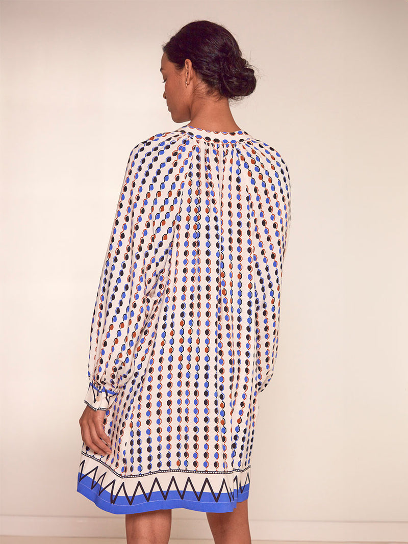 Back View of a Woman Standing Wearing lemlem Meaza Button Up Dress featuring diamond pattern in natural terracotta and rich blue hues against a cream background