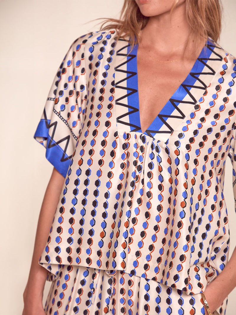 Close up on a Woman Standing Wearing lemlem Rita V Neck Top featuring diamond pattern in natural terracotta and rich blue hues against a cream background and matching Desta Pants