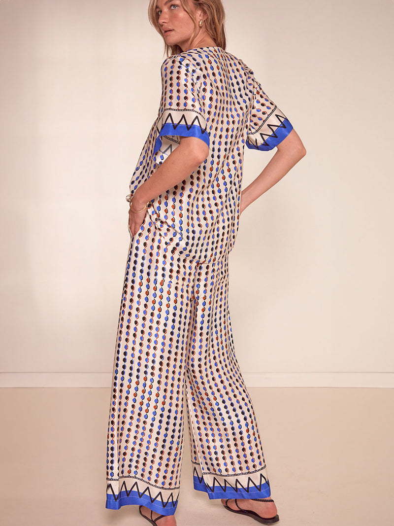 Back of a Woman Standing Wearing lemlem Desta Pants featuring diamond pattern in natural terracotta and rich blue hues against a cream background and matching Rita V Neck Top