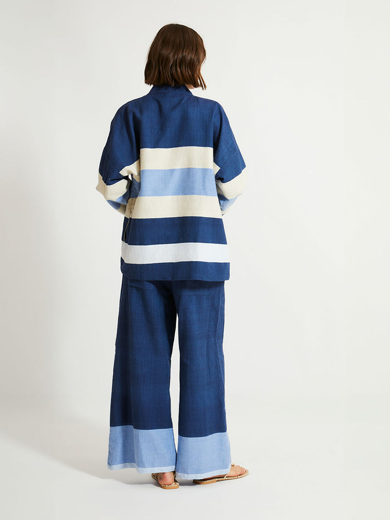 Back of a Woman Standing Wearing lemlem Desta Pants and Terrara Jacket featuring a striking color-block combination of three shades of blue, paired with bands of neutral cream and white, inspired by handmade indigo quilting and African textil.