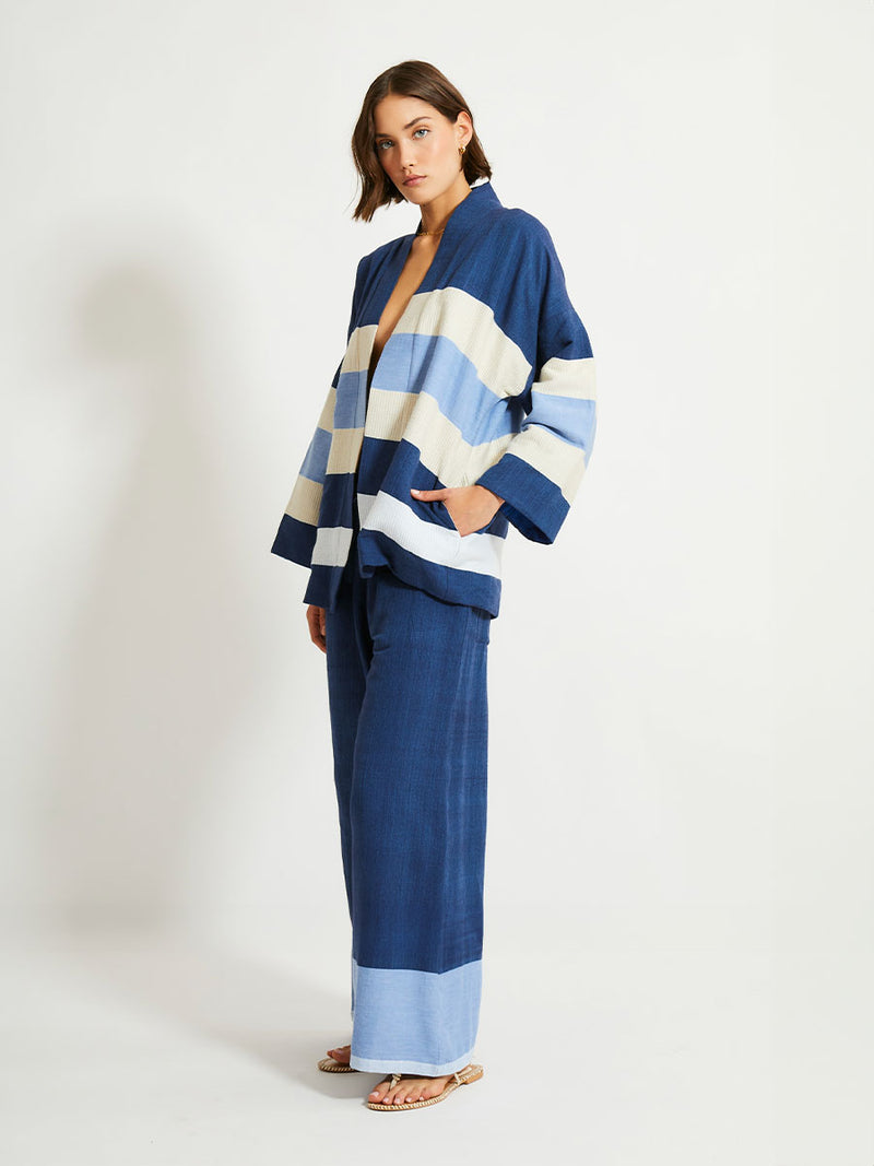 Side of a Woman Standing Wearing lemlem Desta Pants and Terrara Jacket featuring a striking color-block combination of three shades of blue, paired with bands of neutral cream and white, inspired by handmade indigo quilting and African textil.