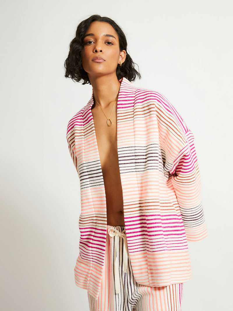 Woman Standing Wearing lemlem Terarra Jacket and matching Wide Leg Pants featuring ombre stripe pattern, where deep plum seamlessly transitions into peach, chestnut, and bright pink