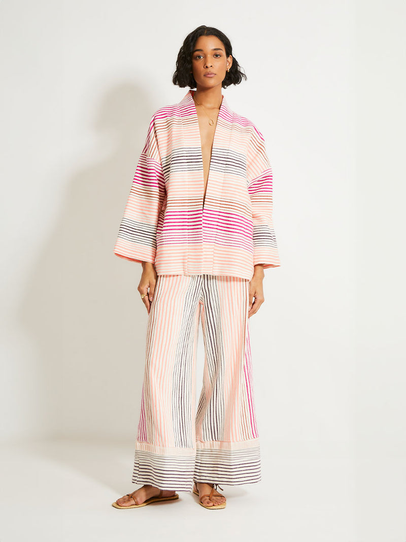 Woman Standing Wearing lemlem Terarra Jacket and matching Wide Leg Pants featuring ombre stripe pattern, where deep plum seamlessly transitions into peach, chestnut, and bright pink