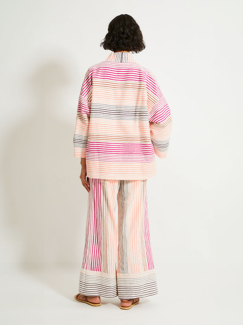 Back of a Woman Standing Wearing lemlem Terarra Jacket and matching Wide Leg Pants featuring ombre stripe pattern, where deep plum seamlessly transitions into peach, chestnut, and bright pink