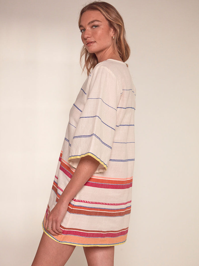 Side View of a Woman Standing wearing lemlem Belkis Caftan featuring tibeb inspired stripes in a vibrant fiesta of colors against a creamy vanilla background