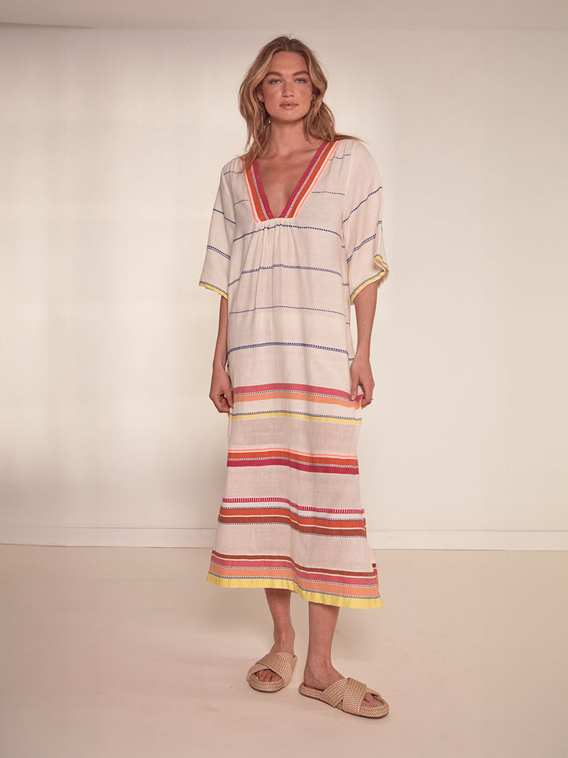 Woman Standing Wearing  lemlem Edna Dress featuring tibeb inspired stripes in a vibrant fiesta of colors against a creamy vanilla background.