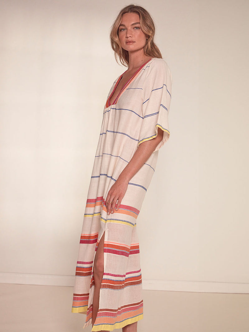 Side View of a Woman Standing Wearing lemlem Edna Dress featuring tibeb inspired stripes in a vibrant fiesta of colors against a creamy vanilla background.