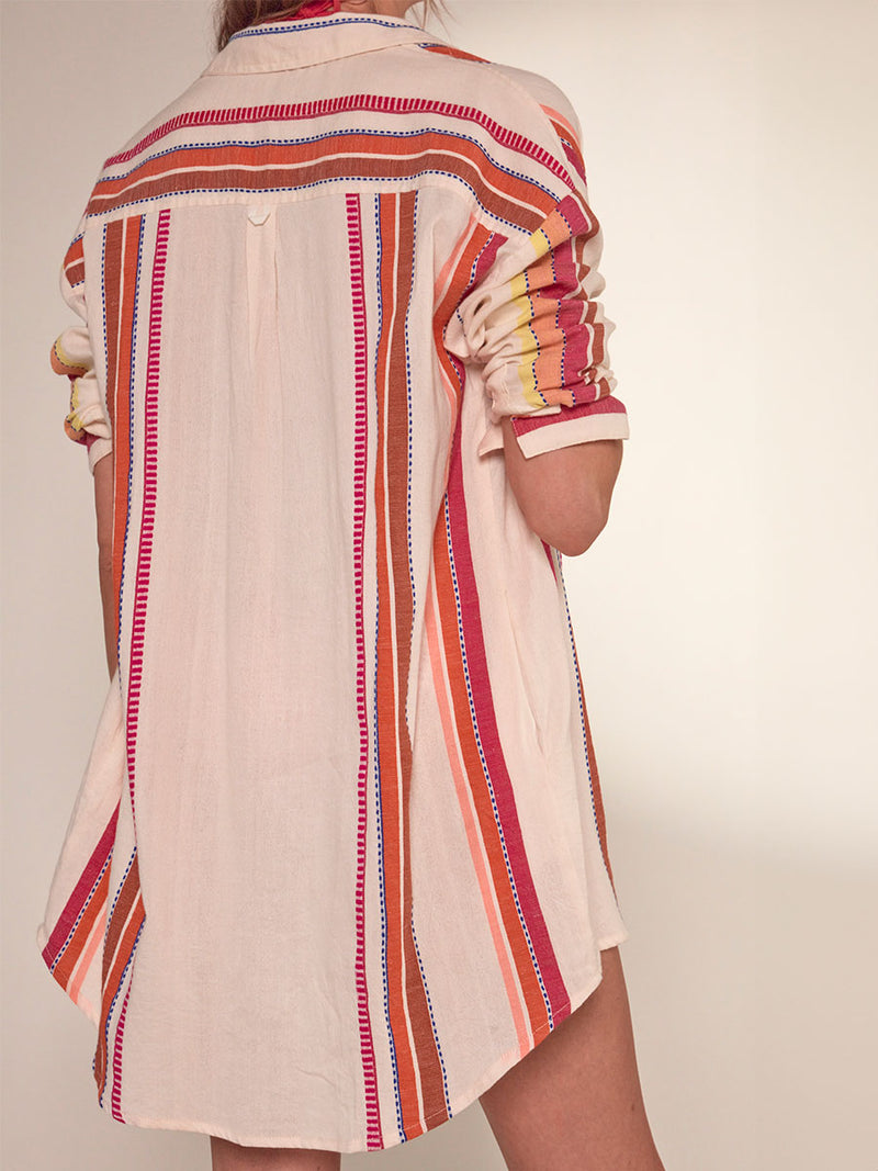 Back View of a woman standing wearing lemlem Mariam Shirt featuring tibeb inspired stripes in a vibrant fiesta of colors against a creamy vanilla background.